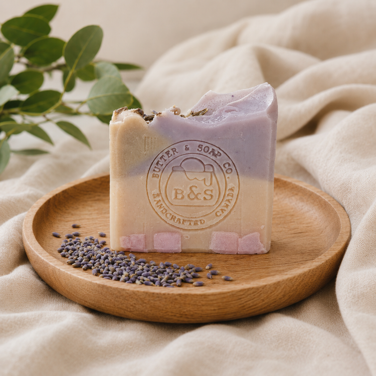 A handmade lavender soap bar with a rustic broken edge sits on a round wooden tray, surrounded by loose lavender buds and green eucalyptus leaves on a beige fabric background. The soap features a circular 'Butter & Soap Co' logo embossed on the front.