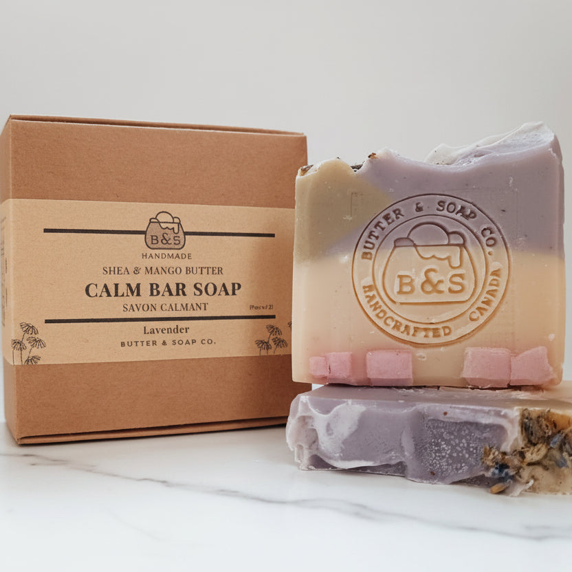 A handmade bar soap set from Butter & Soap Co. featuring a lavender and mango butter layered bar, a smaller pink and cream bar, and a textured herbal bar, displayed next to their kraft paper packaging box.
